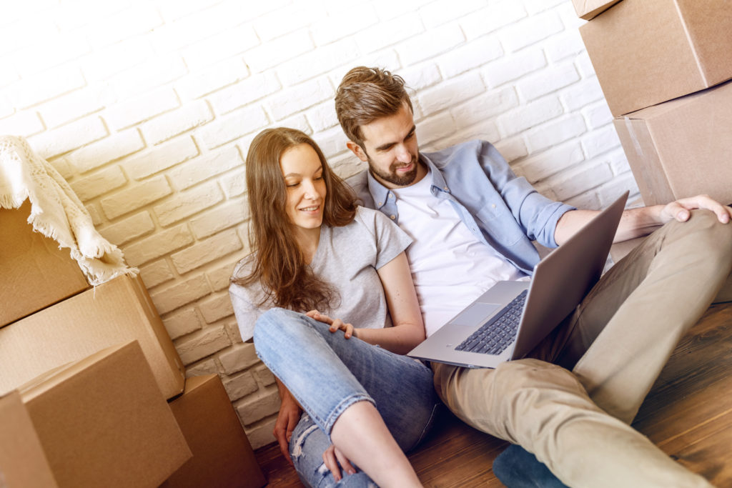 Man and woman sitting in embrace on floor with laptop chatting and moving to new flat.